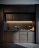 A modern kitchen with dark cabinetry and a sleek, minimalist design. The countertop is made of dark marble, and there's a built-in stove. Under-cabinet lighting provides a warm glow. A knife block and a couple of cutting boards are placed on the counter next to the stove.