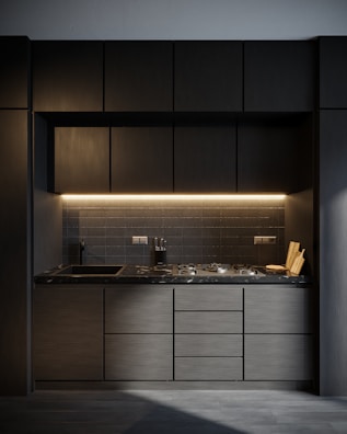 A modern kitchen with dark cabinetry and a sleek, minimalist design. The countertop is made of dark marble, and there's a built-in stove. Under-cabinet lighting provides a warm glow. A knife block and a couple of cutting boards are placed on the counter next to the stove.