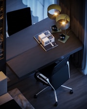 A modern workspace featuring a sleek, dark-colored desk with books and stylish lamps. A black ergonomic office chair is positioned at the desk, which is set against a backdrop of contemporary decor, including curtains and flooring.