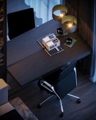 A modern workspace featuring a sleek, dark-colored desk with books and stylish lamps. A black ergonomic office chair is positioned at the desk, which is set against a backdrop of contemporary decor, including curtains and flooring.