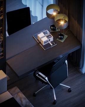 A modern workspace featuring a sleek, dark-colored desk with books and stylish lamps. A black ergonomic office chair is positioned at the desk, which is set against a backdrop of contemporary decor, including curtains and flooring.