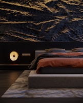 A modern bedroom featuring a textured dark wall with wave-like patterns. The bed has a low-profile design, dressed in dark gray bedding with a burnt orange blanket. A contemporary round lamp with a soft glow sits on a sleek bedside table. The room is minimalistic, with a sense of balance and harmony.