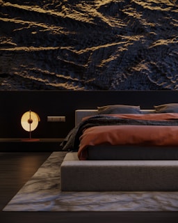 A modern bedroom featuring a textured dark wall with wave-like patterns. The bed has a low-profile design, dressed in dark gray bedding with a burnt orange blanket. A contemporary round lamp with a soft glow sits on a sleek bedside table. The room is minimalistic, with a sense of balance and harmony.