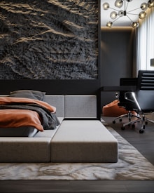 A modern, minimalist bedroom with a low-profile gray bed and an elegant textured wall that appears to be inspired by natural stone formations. The bed is adorned with neatly arranged pillows and a blanket in muted orange and dark gray hues. To the side, there's a black office chair on wheels next to a sleek, dark-colored desk, positioned near a window with sheer curtains. A unique chandelier with multiple lights hangs above, adding a contemporary touch to the space.