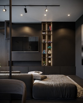 A modern, dimly-lit living room with a minimalist design, featuring a large black flat-screen TV mounted on a dark wall, under subtle recessed lighting. A sleek shelving unit beside the TV holds several books and decorative items, including a vase with blue flowers. A comfortable gray sectional sofa and a small round table with a book are positioned in the foreground. The color palette includes dark tones and soft textures, enhancing the sophisticated atmosphere.