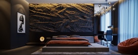 A modern bedroom features a large textured wall behind a minimalist bed. The room is decorated with warm lighting and a sophisticated hanging light fixture. A work desk and chair are situated by tall windows with sheer curtains, and a contemporary art piece hangs on one wall. A bean bag sits in one corner.