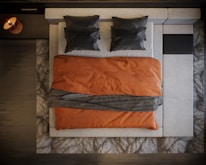 A neatly arranged bed viewed from above, featuring two dark pillows on a light-colored mattress. An orange blanket covers the bed, with a dark textured throw draped across the middle. The bed is framed by a patterned rug and dark wooden floor.