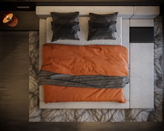 A neatly arranged bed viewed from above, featuring two dark pillows on a light-colored mattress. An orange blanket covers the bed, with a dark textured throw draped across the middle. The bed is framed by a patterned rug and dark wooden floor.