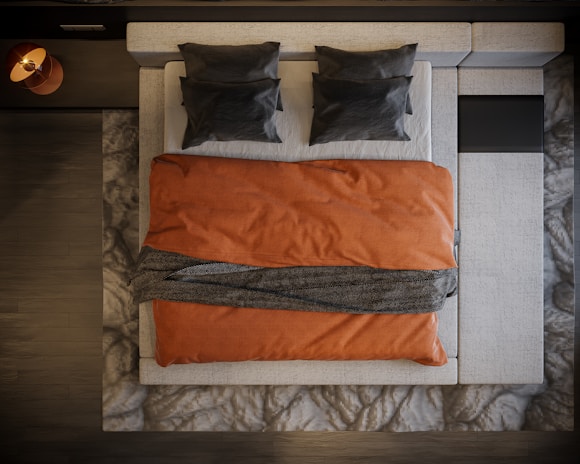 A neatly arranged bed viewed from above, featuring two dark pillows on a light-colored mattress. An orange blanket covers the bed, with a dark textured throw draped across the middle. The bed is framed by a patterned rug and dark wooden floor.