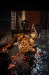 A large piece of meat is being roasted over an open flame with glowing embers and smoke rising. The cooking area has a rustic aesthetic, featuring brick walls and wooden elements with visible metal fixtures in the background.
