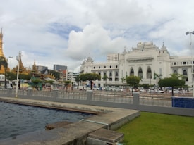 A grand white colonial-style building with intricate architectural details stands prominently. In the foreground, a body of water and a grassy area are visible, separated by a low fence. To the left, golden pagodas with spires can be seen, contrasting with a backdrop of modern buildings. Cars and a bus are visible on the road in front of the central building.