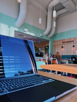 A modern cafe interior with a clean and minimalistic design. A laptop is open on a wooden table in the foreground, displaying a screen with text. The cafe features high ceilings, pastel green and wood paneling, with large air ducts visible. In the background, people are seated on colorful chairs at tables, engaged in conversation and working.