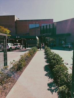 A pathway lined with bushes leads to a large brick building with glass windows. Several people are walking along the path towards the building entrance. Parked cars are visible to the left near what appears to be a parking area.
