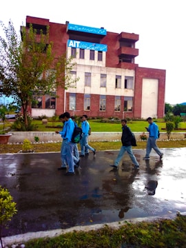 A group of students in blue uniforms walk along a wet pathway in front of a red brick building with large windows. The environment is lush with greenery, including trees and shrubs. The sky is overcast, suggesting recent rain.