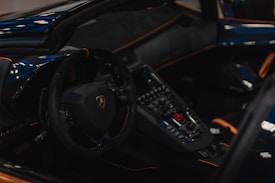 The interior of a luxury sports car features a sleek steering wheel with a prominent logo and an array of control buttons on the dashboard. The design includes glossy black and orange accents with a sophisticated and modern appearance.