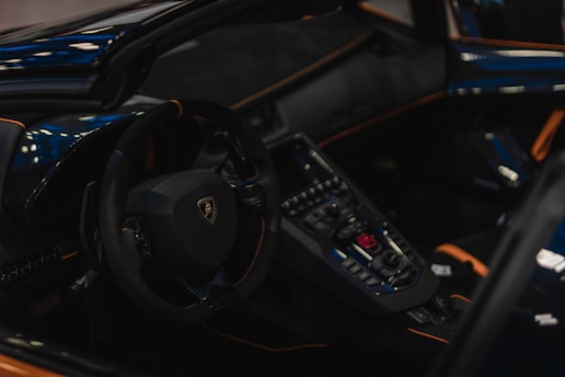 The interior of a luxury sports car features a sleek steering wheel with a prominent logo and an array of control buttons on the dashboard. The design includes glossy black and orange accents with a sophisticated and modern appearance.
