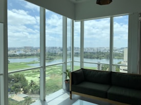 The image features a spacious room with large glass windows offering a panoramic view of a city. Outside, there is a landscape with a river, lush greenery, and numerous buildings in the background, under a partly cloudy sky. Inside the room, there is a dark green sofa and a small potted plant, suggesting a modern, comfortable interior space.