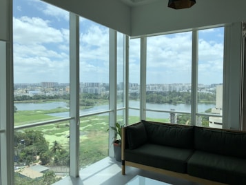 The image features a spacious room with large glass windows offering a panoramic view of a city. Outside, there is a landscape with a river, lush greenery, and numerous buildings in the background, under a partly cloudy sky. Inside the room, there is a dark green sofa and a small potted plant, suggesting a modern, comfortable interior space.