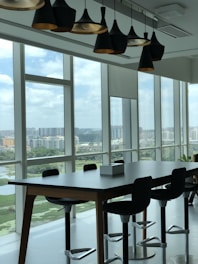 A modern office space with large floor-to-ceiling windows revealing a cityscape. The room features a long wooden table with several high-backed swivel chairs arranged around it. Above the table, a series of contemporary pendant lights hang from the ceiling. The windows offer a view of multiple residential buildings and green areas under a cloudy sky.