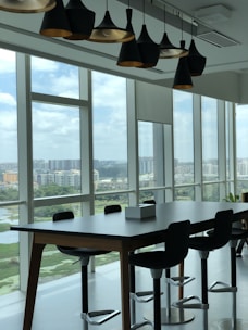 A modern office space with large floor-to-ceiling windows revealing a cityscape. The room features a long wooden table with several high-backed swivel chairs arranged around it. Above the table, a series of contemporary pendant lights hang from the ceiling. The windows offer a view of multiple residential buildings and green areas under a cloudy sky.