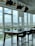 A modern office space with large floor-to-ceiling windows revealing a cityscape. The room features a long wooden table with several high-backed swivel chairs arranged around it. Above the table, a series of contemporary pendant lights hang from the ceiling. The windows offer a view of multiple residential buildings and green areas under a cloudy sky.