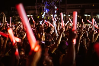 A large crowd of people, mostly young adults, is gathered at a nighttime event, holding glowing red and white light sticks. The atmosphere appears lively and energetic, suggesting a concert or festival setting.