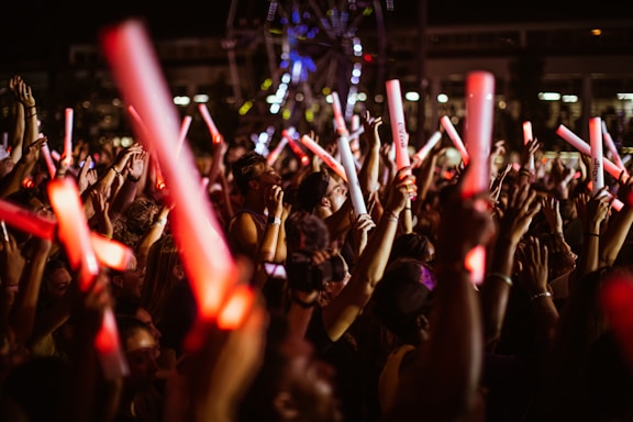 A large crowd of people, mostly young adults, is gathered at a nighttime event, holding glowing red and white light sticks. The atmosphere appears lively and energetic, suggesting a concert or festival setting.