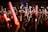 A large crowd of people, mostly young adults, is gathered at a nighttime event, holding glowing red and white light sticks. The atmosphere appears lively and energetic, suggesting a concert or festival setting.