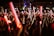 A large crowd of people, mostly young adults, is gathered at a nighttime event, holding glowing red and white light sticks. The atmosphere appears lively and energetic, suggesting a concert or festival setting.