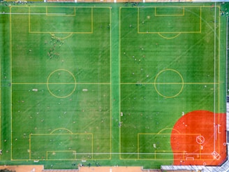 Aerial view of two adjacent soccer fields with clearly marked boundaries and goalposts. Players are scattered across both fields, with some gathered in small groups, suggesting either practice or multiple games occurring simultaneously. The fields are bordered by fences, with an additional area featuring a reddish-colored surface, possibly a baseball field.