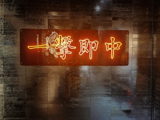 A neon sign with Chinese characters is mounted on a textured wall. The sign emits a warm, glowing light, creating a contrast against the darker, metallic background.