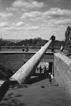 A large telescope structure is mounted on a brick platform overlooking a paved walkway. People are walking along the path, and some are positioned at the observatory. The sky is filled with scattered clouds, and the city skyline is visible in the background. The telescope is covered with graffiti.