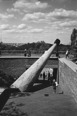 A large telescope structure is mounted on a brick platform overlooking a paved walkway. People are walking along the path, and some are positioned at the observatory. The sky is filled with scattered clouds, and the city skyline is visible in the background. The telescope is covered with graffiti.