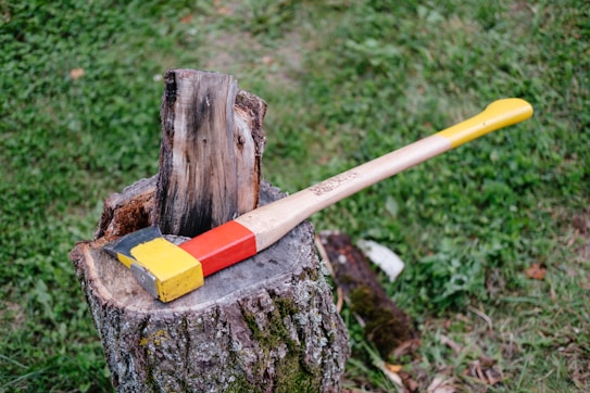 An axe with a brightly colored handle featuring yellow and red sections is placed on a tree stump. The stump shows signs of prior use, with a piece of wood partially split on top. The setting is outdoors, with green grass visible in the background.