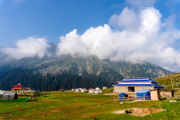 A picturesque mountainous landscape featuring lush green grass, scattered colorful tents, and a traditional building with a blue roof. Towering mountains in the background are partially obscured by fluffy white clouds, creating a serene and tranquil atmosphere.