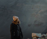 Model wearing layered rugged outdoor gear against a rocky mountain backdrop at dawn.
