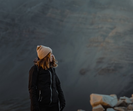 A person in outdoor gear stands on a rocky surface, wearing a beige beanie and a dark jacket. The background features a vast, rugged mountain or rocky terrain with muted colors.