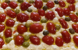 A close-up view of a focaccia bread topped with cherry tomatoes, green olives, and melted cheese. The ingredients are arranged in a pattern, with the vibrant red of the tomatoes contrasting against the green olives and creamy white cheese. Olive oil glistens on the surface, adding a rich, golden hue.