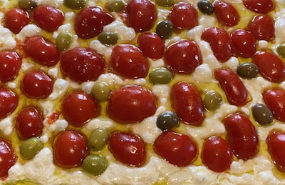 A close-up view of a focaccia bread topped with cherry tomatoes, green olives, and melted cheese. The ingredients are arranged in a pattern, with the vibrant red of the tomatoes contrasting against the green olives and creamy white cheese. Olive oil glistens on the surface, adding a rich, golden hue.