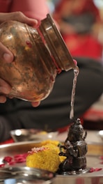 A person pours water from a copper vessel carefully onto a small, intricately carved metallic statue. The setting includes marigold flowers, petals, and a stainless steel tray. The background features blurred colors, predominantly red and black, adding a vibrant contrast to the foreground.