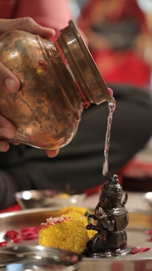 A person pours water from a copper vessel carefully onto a small, intricately carved metallic statue. The setting includes marigold flowers, petals, and a stainless steel tray. The background features blurred colors, predominantly red and black, adding a vibrant contrast to the foreground.