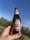 A bottle of Peroni beer is held up against a clear blue sky. The label is prominently displayed with its distinctive red and gold design featuring the brand name and Italian flag colors. The background includes a blurred view of trees and grass, suggesting an outdoor setting.