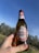 A bottle of Peroni beer is held up against a clear blue sky. The label is prominently displayed with its distinctive red and gold design featuring the brand name and Italian flag colors. The background includes a blurred view of trees and grass, suggesting an outdoor setting.