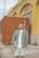 A person stands holding a guitar in an urban setting. They are wearing a green jacket over a white shirt and shorts. Behind them is a large orange shipping container and a yellow building with peeling paint.