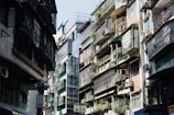 An urban street scene featuring closely-packed residential buildings. Apartments have metal railings and are adorned with potted plants, with air conditioning units attached to many balconies. Sunlight casts shadows, creating a contrast between the bright and dark areas.