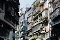 An urban street scene featuring closely-packed residential buildings. Apartments have metal railings and are adorned with potted plants, with air conditioning units attached to many balconies. Sunlight casts shadows, creating a contrast between the bright and dark areas.