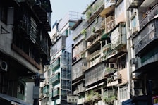 An urban street scene featuring closely-packed residential buildings. Apartments have metal railings and are adorned with potted plants, with air conditioning units attached to many balconies. Sunlight casts shadows, creating a contrast between the bright and dark areas.