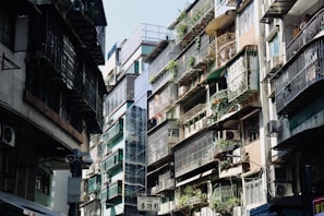 An urban street scene featuring closely-packed residential buildings. Apartments have metal railings and are adorned with potted plants, with air conditioning units attached to many balconies. Sunlight casts shadows, creating a contrast between the bright and dark areas.