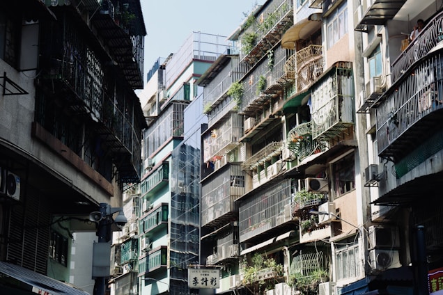 An urban street scene featuring closely-packed residential buildings. Apartments have metal railings and are adorned with potted plants, with air conditioning units attached to many balconies. Sunlight casts shadows, creating a contrast between the bright and dark areas.