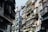 An urban street scene featuring closely-packed residential buildings. Apartments have metal railings and are adorned with potted plants, with air conditioning units attached to many balconies. Sunlight casts shadows, creating a contrast between the bright and dark areas.